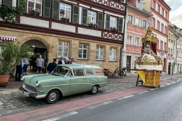 Außenansicht der Metzgerei Hornung in Bamberg mit Oldtimer vor dem Fachwerk. Rechts steht die Fronleichnams-Figur zur Prozession bereit.