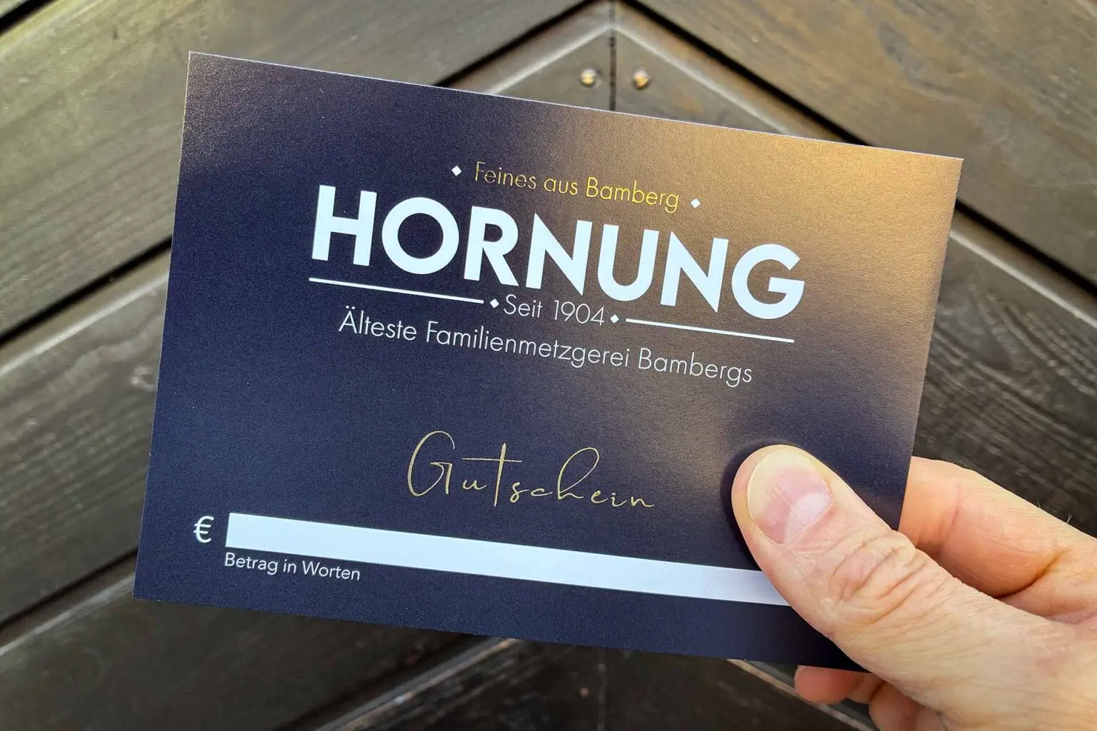 Gutschein der Metzgerei Hornung in Bamberg vor Holztür gehalten