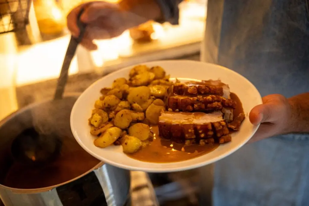 Mitarbeiter schöpft Bratensoße aus einem Topf und gießt es über den Krustenbraten mit Bratkartoffeln