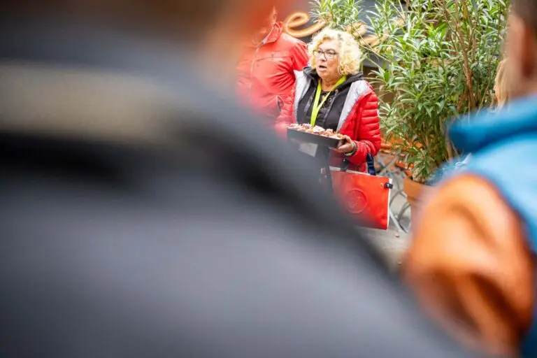 Frau mit blondem, lockigen Haar und Brill in roter Jacke, hält eine Schale mit Kostproben bereit und erklärt den Teilnehmern der Genussführung etwas zur Metzgerei Hornung